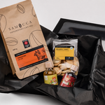 Schwarze Geschenkbox mit schwarzem Seidenpapier und einer Auswahl an Samocca-Produkten. Enthalten sind ein Beutel Kaffee aus Kenia, französische Mandel-Sablé-Butterkekse sowie zwei kleine Flaschen mit Sirup. Die Produkte sind dekorativ in der geöffneten Geschenkbox angeordnet. Die sommerliche Variante des Geschenkkorbs kommt ohne Schokolade aus.