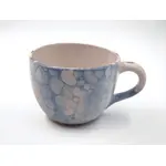 Keramiktasse mit Henkel in Blau- und Weißtönen mit marmorierter Oberfläche steht auf einer hellen Fläche vor neutralem Hintergrund.
