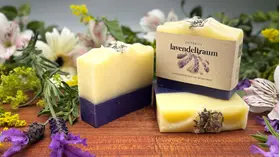 Drei Seifen liegen auf einem Holzbrett. Die Seifen sind lila weiß. Im Hintergrund sind bunte Blumen zu sehen.