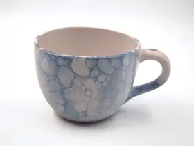 Keramiktasse mit Henkel in Blau- und Weißtönen mit marmorierter Oberfläche steht auf einer hellen Fläche vor neutralem Hintergrund.
