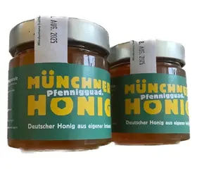 Zwei Gläser mit einer gelben Aufschrift Münchner Pfennig guad Honig und grünem Etikett, gefüllt mit dunkel gelbem Honig.