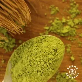 Grünes Matchapulver auf Esslöffel. Hintergrund in Holzoptik und ein Matchabesen mit zerstreutem Matchapulver.