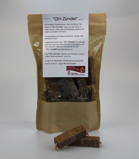 Braune, wiederverschließbare Verpackung mit der Aufschrift Oh!-Zünder, gefüllt mit mehreren dunklen Anzündrechtecken; davor liegen zwei einzelne Rechtecke mit sichtbarem Docht auf weißem Hintergrund.